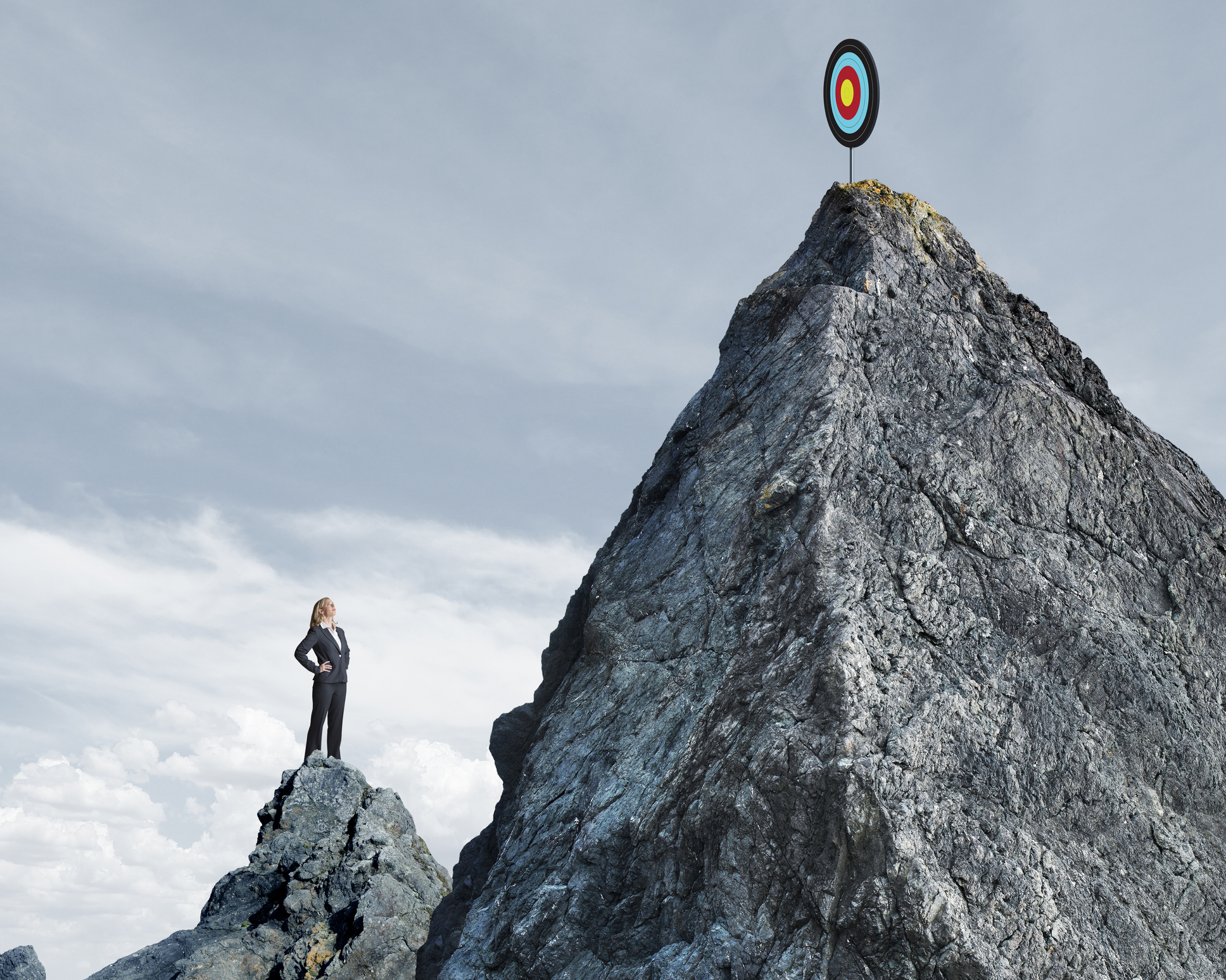 A businesswoman stands on top of a small mountain and looks up at a target that sits on top of a taller mountain top that rises in front of her.